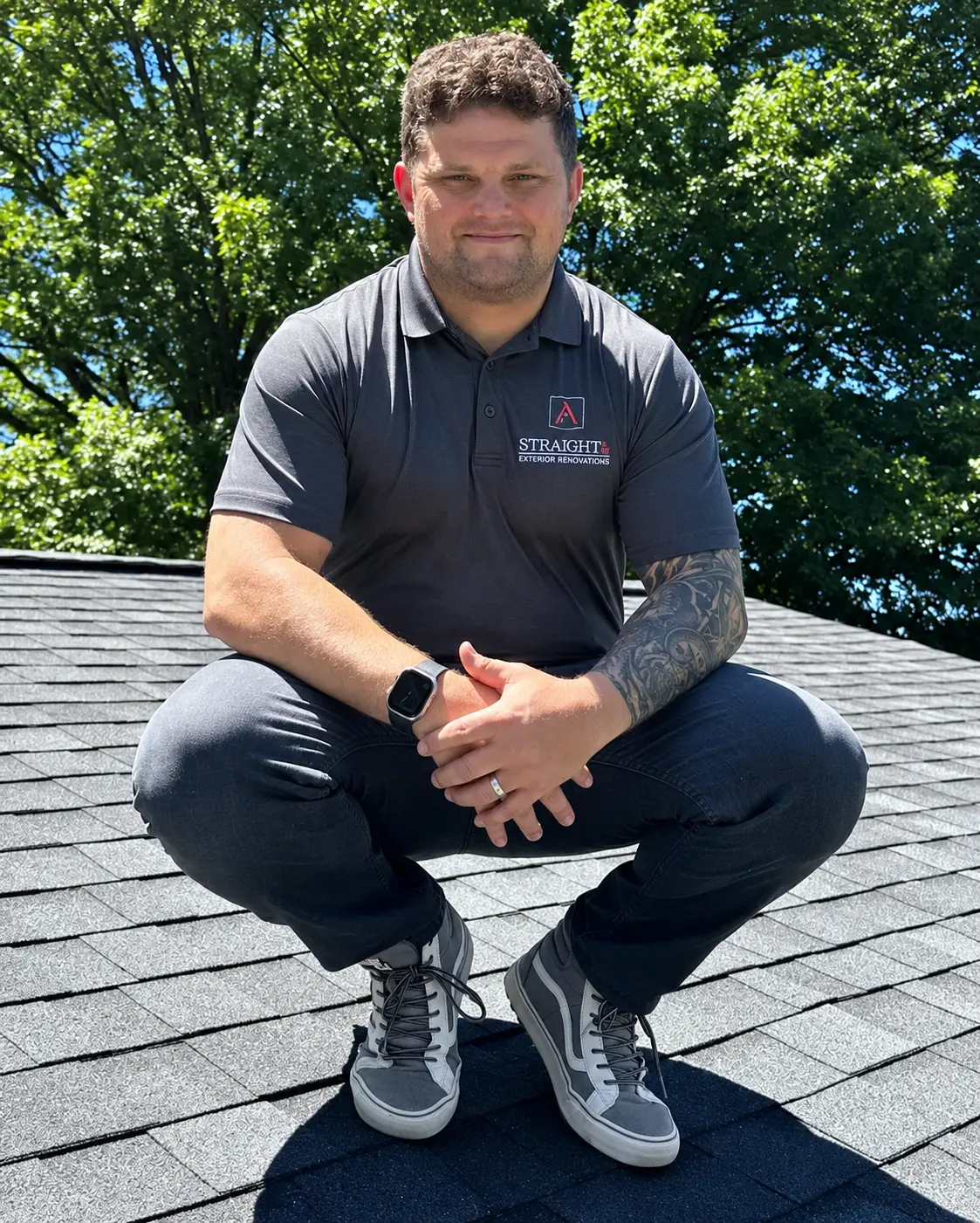 Matt Alexander, owner of Straight A Exterior Renovations, crouching on a shingle roof in a company polo
